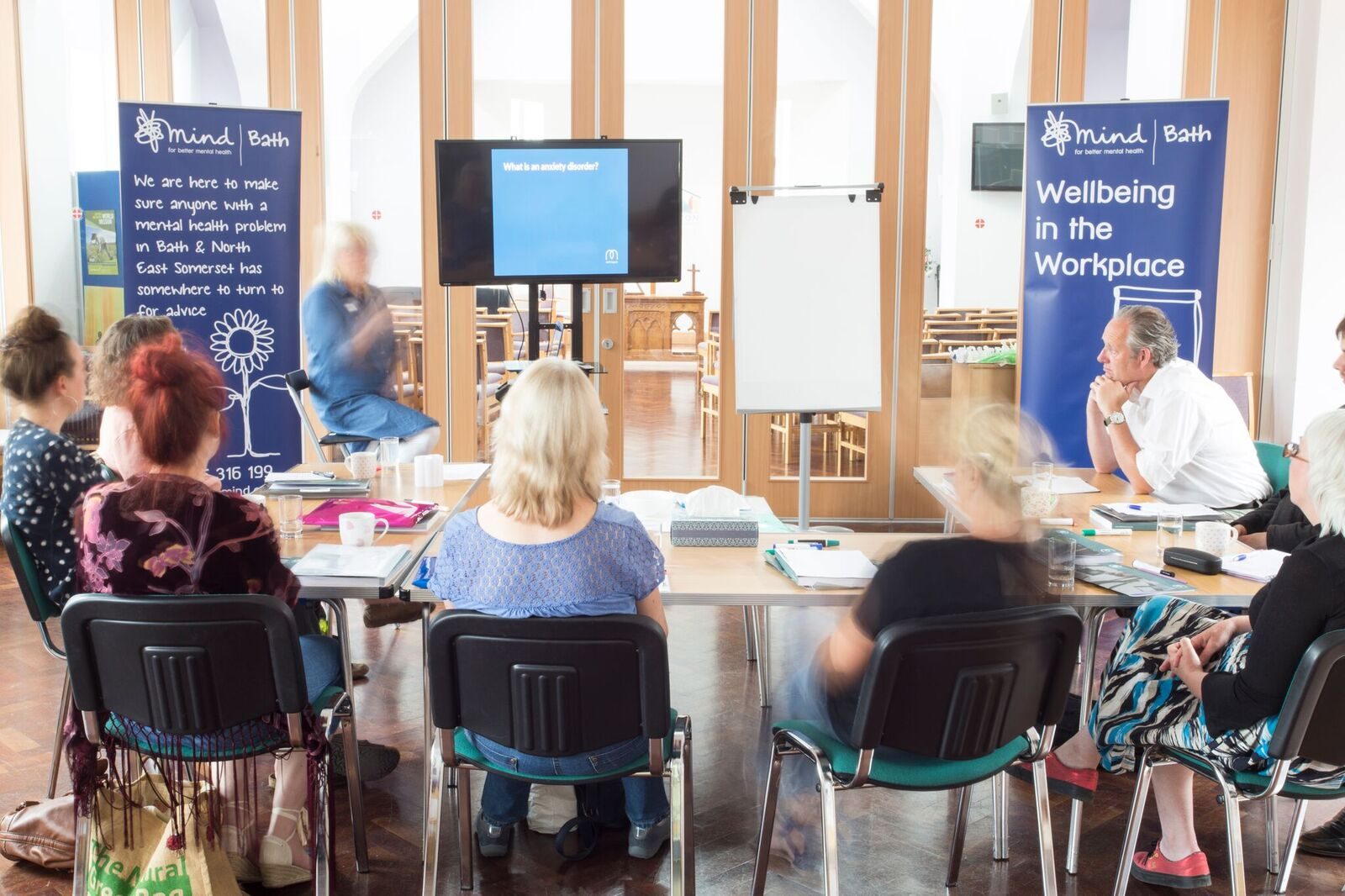 A group of people sitting on chairs around a table with banners that say Wellbeing in the Workplace