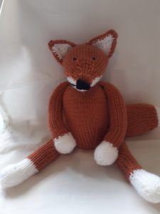 A brown knitted fox with white hands and feet sits against a white background. 
