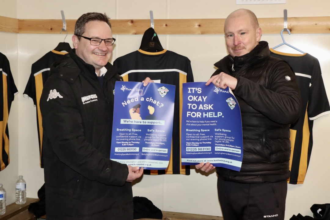 Two white men holding a poster in a football changing room. The poster advertises the charity Bath Mind's mental health support services