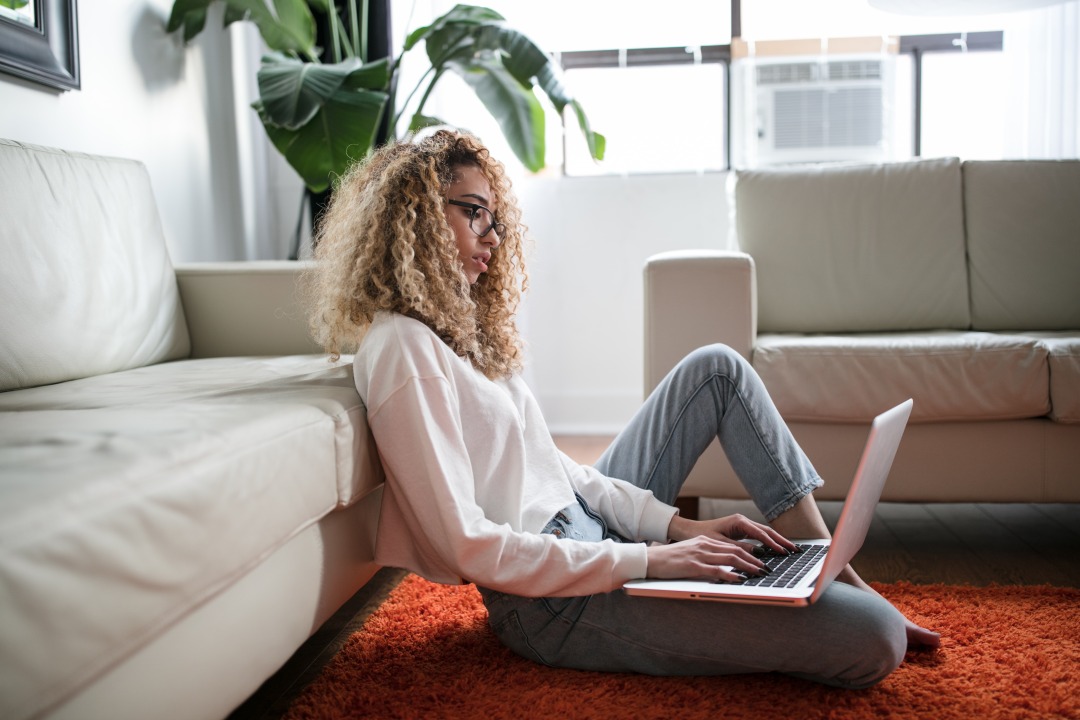 A woman sits on the floor by a sofa on her laptop.