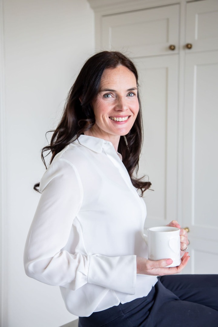 A white woman sitting on a chair holding a mug. She has long brown hair and is smiling inti the camera.