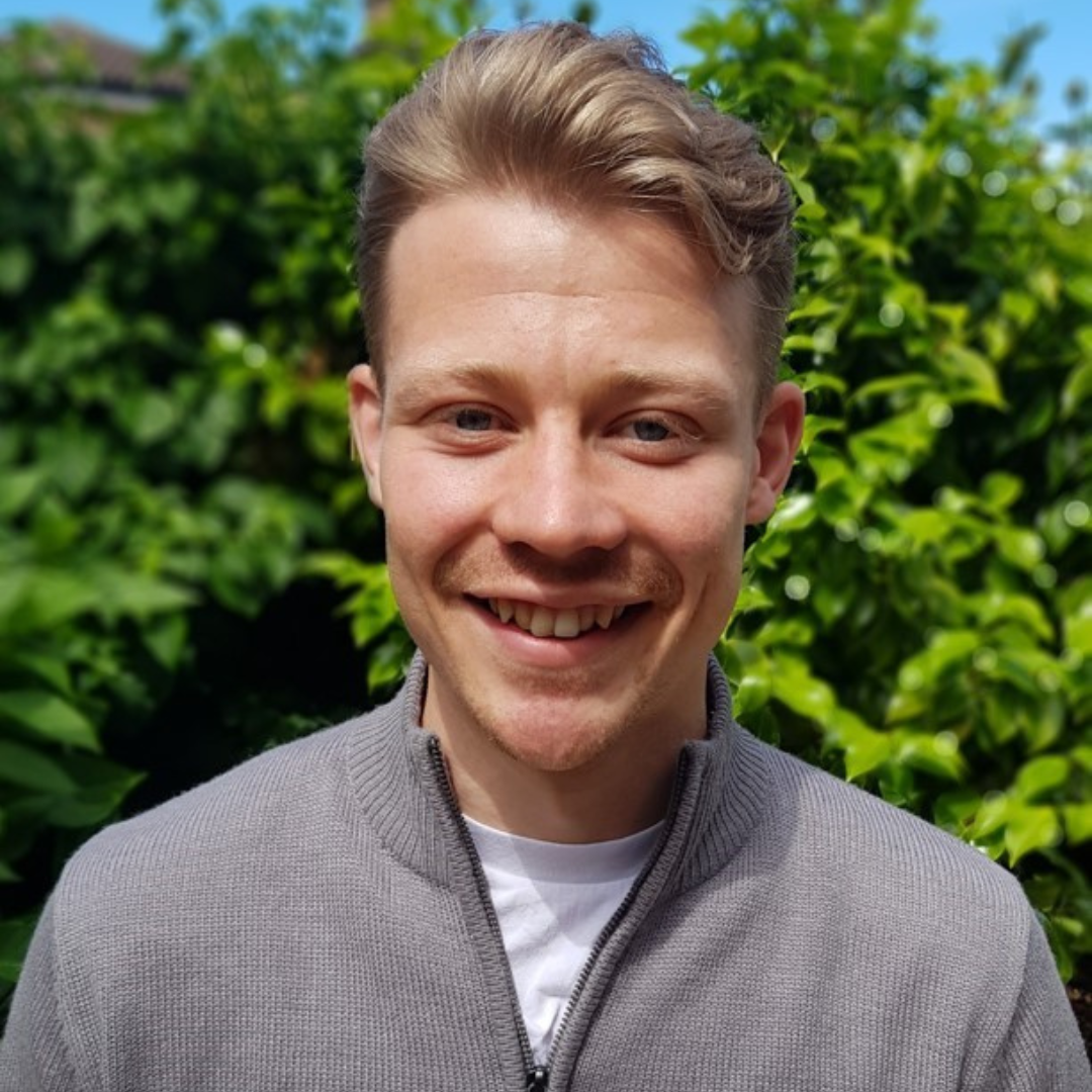 Portrait of a white man with blonde hair stood in front of a green bush. He smiles at the camera.