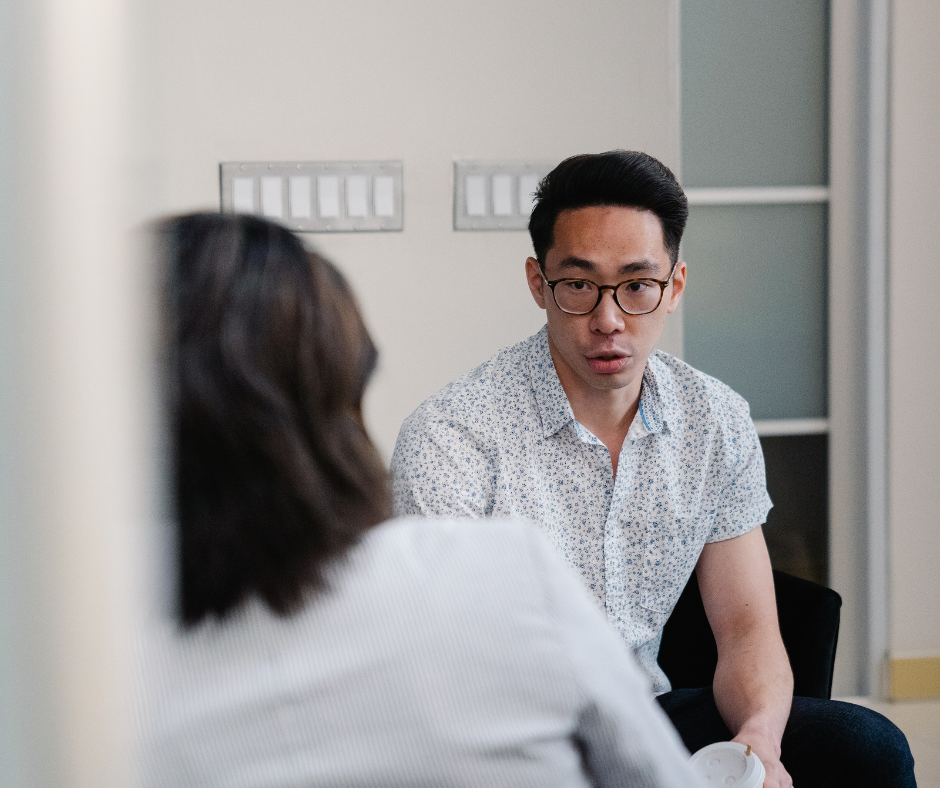 A man with glasses wearing a white shirt is sat on a sofa opposite another person. He likes like he's talking and very thoughtful.