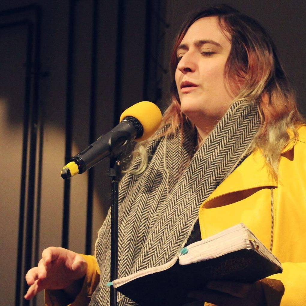 A white woman speaks into a microphone. She has brown hair and is wearing a yellow jacket. She holds a notebook in one hand.