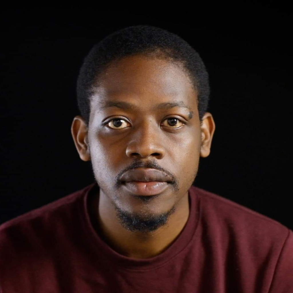 A portrait of a black man wearing a dark red jumper. He has short hair and trimmed facial hair.