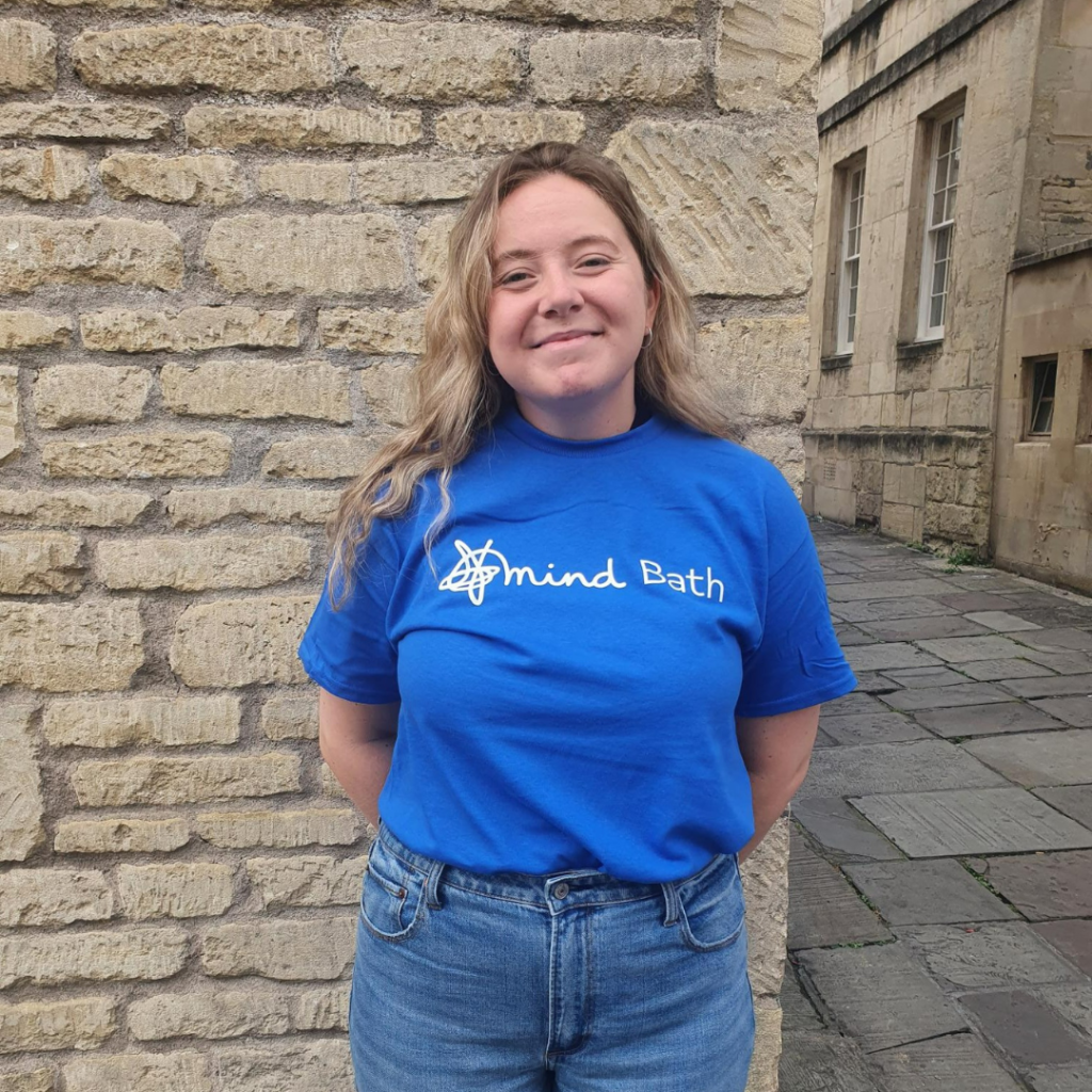 A white girl with blonde hair stands against a wall. She is smiling and wearing a blue t-shirt that says Bath Mind on it