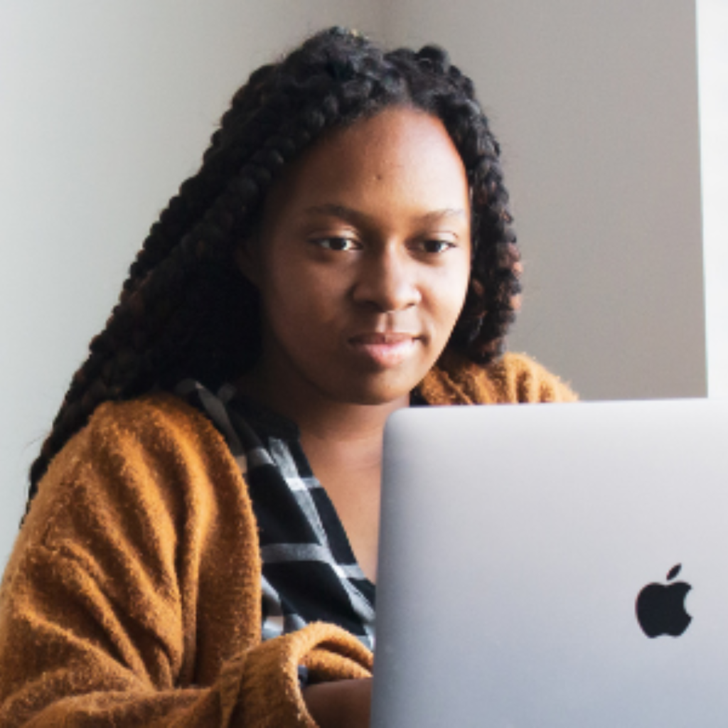 A black girl sits in front of her open laptop. She wears a brown cardigan and looks content