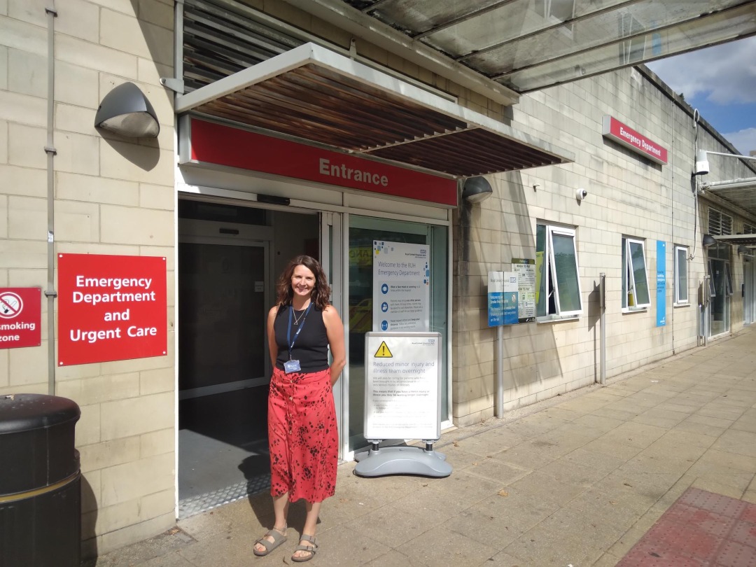 A white woman with brown hair wearing a red skirt and black top stands in front of the Emergency Department at a hospital. She has her hands behind her back and is smiling.