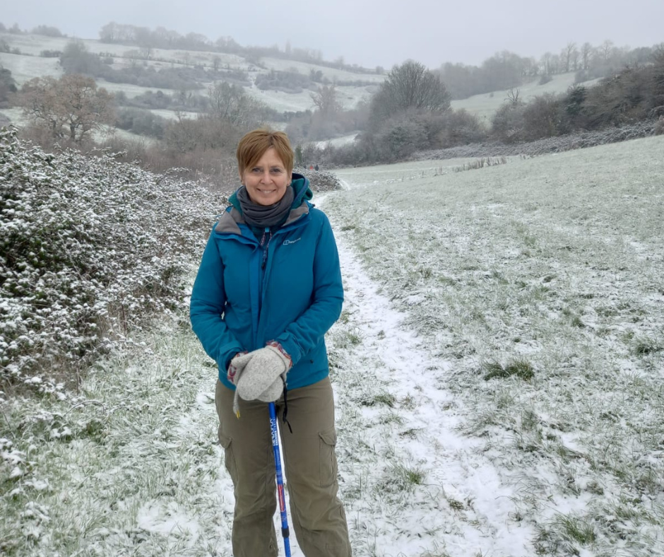 A white woman stands in a snowy field. She has short red hair and wear a green waterproof coat and brown trousers. She is holding a walking pole.