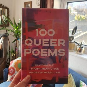 A hand holds 100 Queer Poems book up to a sunny window.