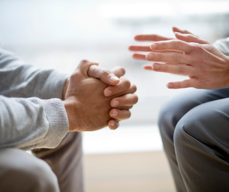 Two people sit across from each other. We see their hands and it seems as though they are in a discussion.