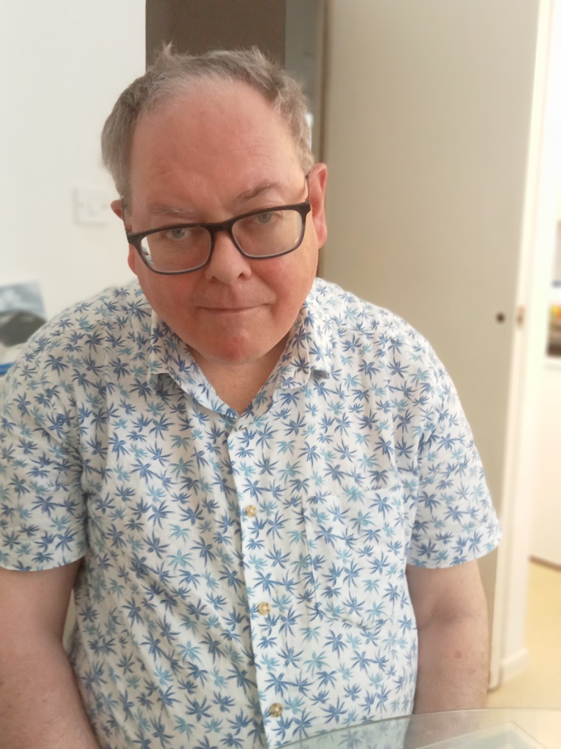 A white man wearing glasses and a floral shirt is sat on a chair looking into the camera.