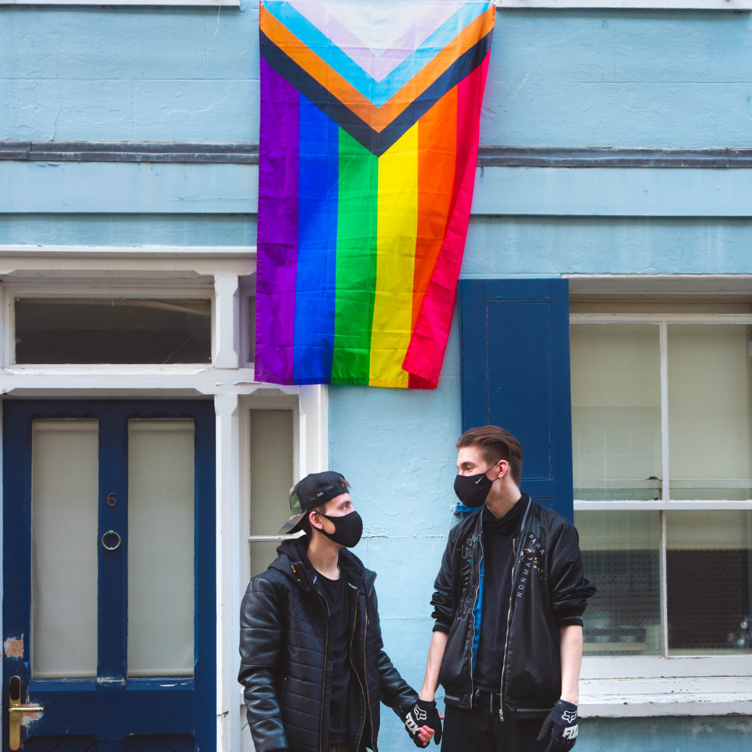 Two people stand looking at each other holding hands. They were black face masks and are stood in front of the pride flag.