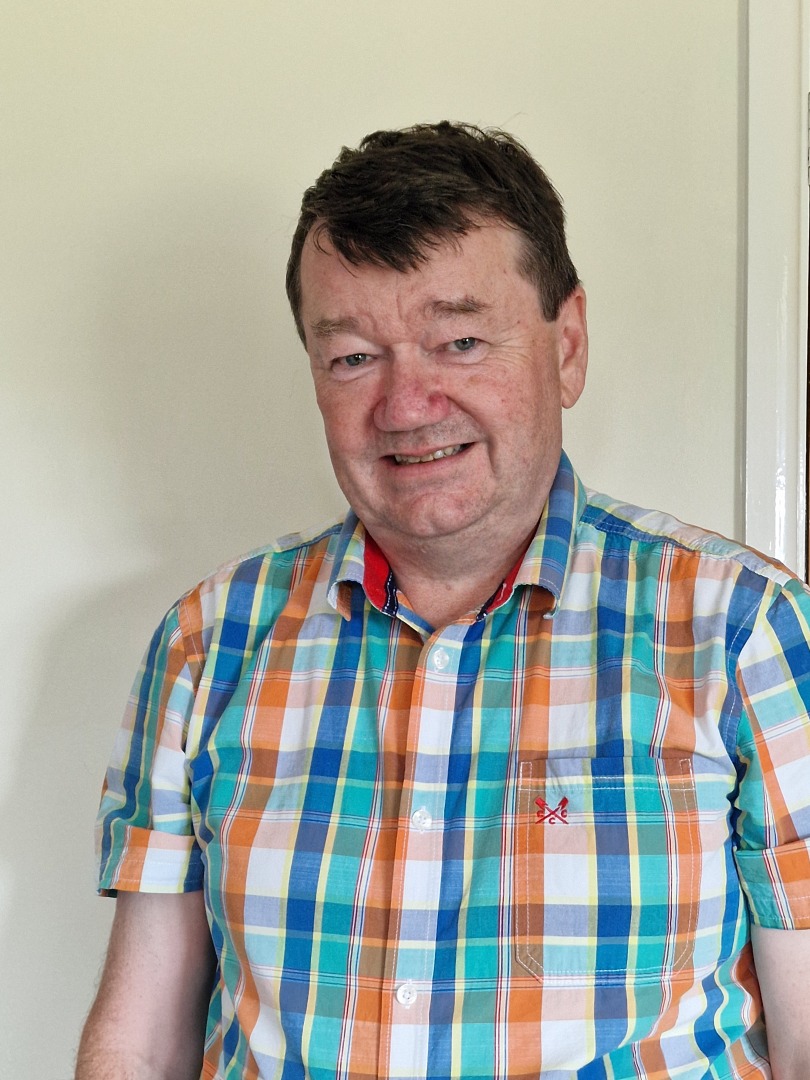 A portrait photo of a white man with brown hair wearing a check shirt.