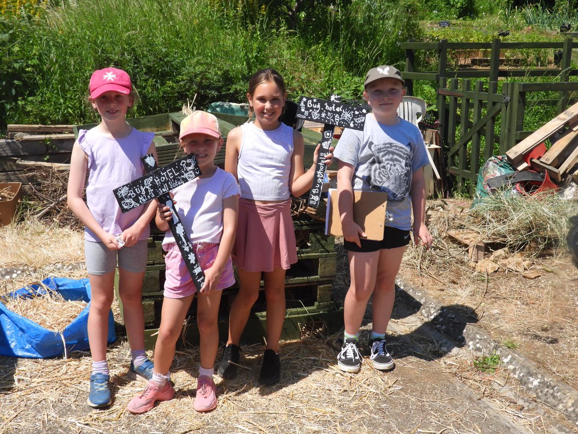 A group of school children are stood in some allotments carrying a bug hotel sign.