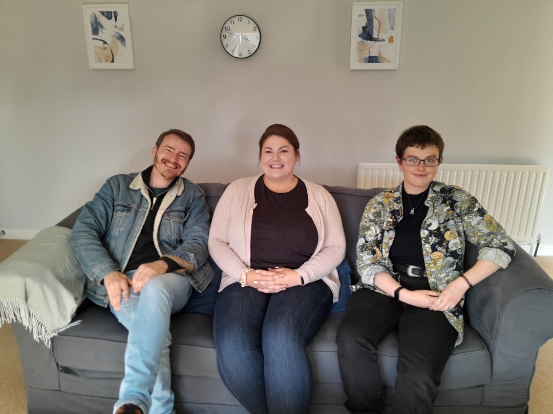 Three people sit on a sofa together smiling.