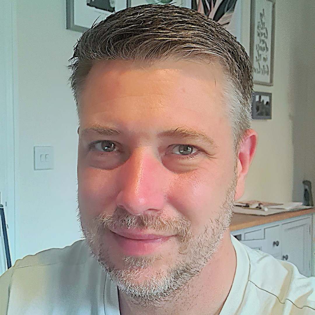 A white man with short hair and facial hair smiles into the camera