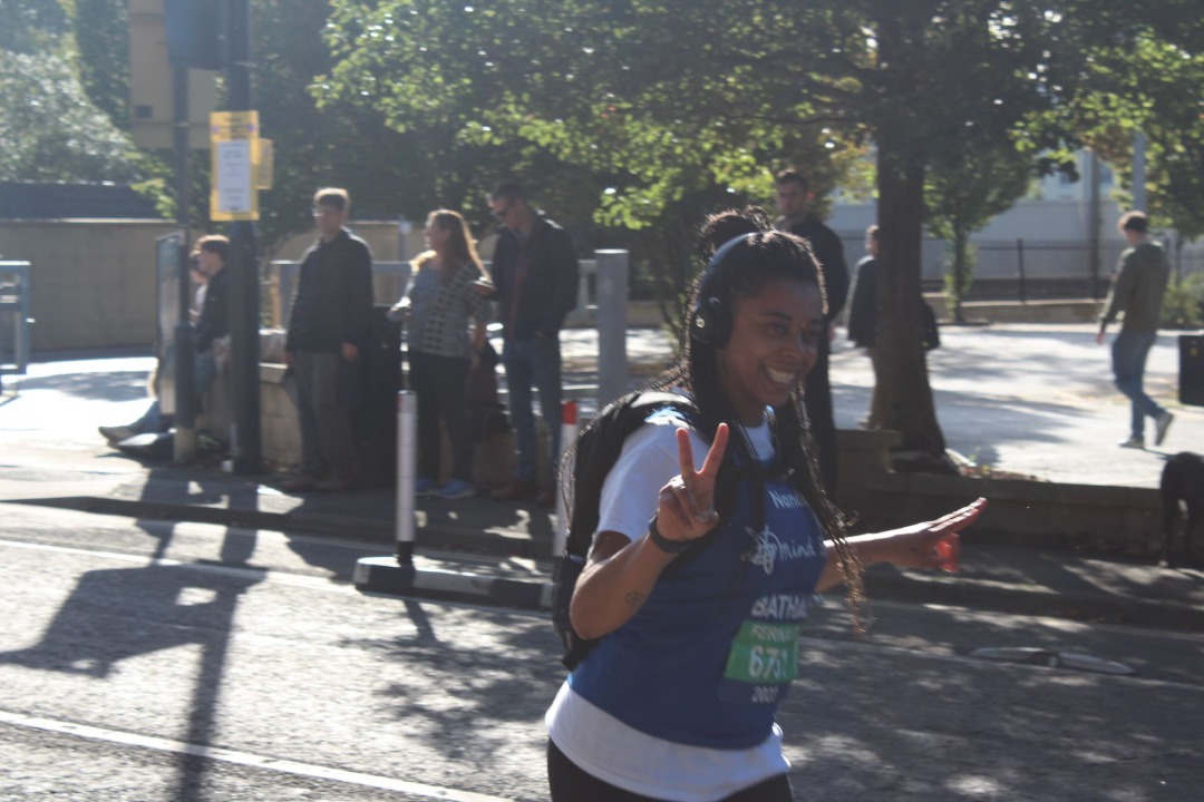 A black woman with headphones on and wearing a Bath Mind blue vest smiles and does the peace sign as she runs.