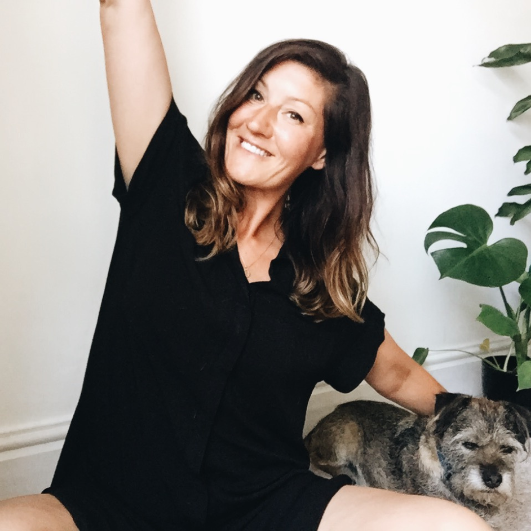 A white woman with brown hair stretches while sat on the floor by a dog.