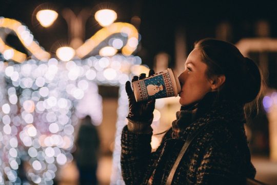 A woman drinks from a takeaway coffee cup while staring at Christmas lights and decorations