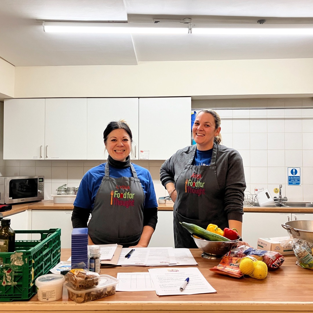 Two women stand in a kitchen wearing Food for Thought aprons