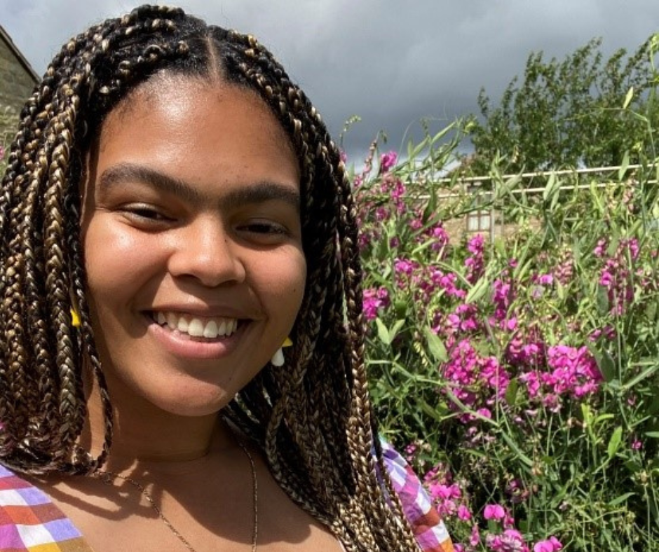 A black women wears a multicoloured dress and stands in a flower filled field smiling.