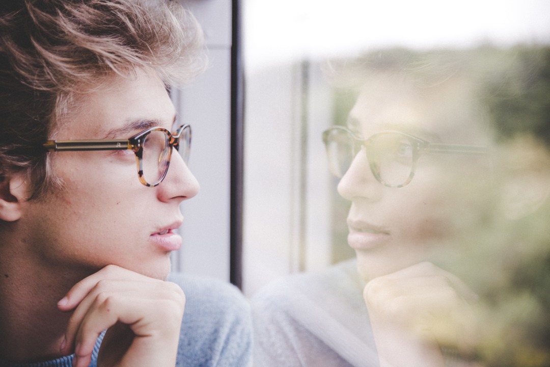 A white man with blonde hair in glasses stares out of a window