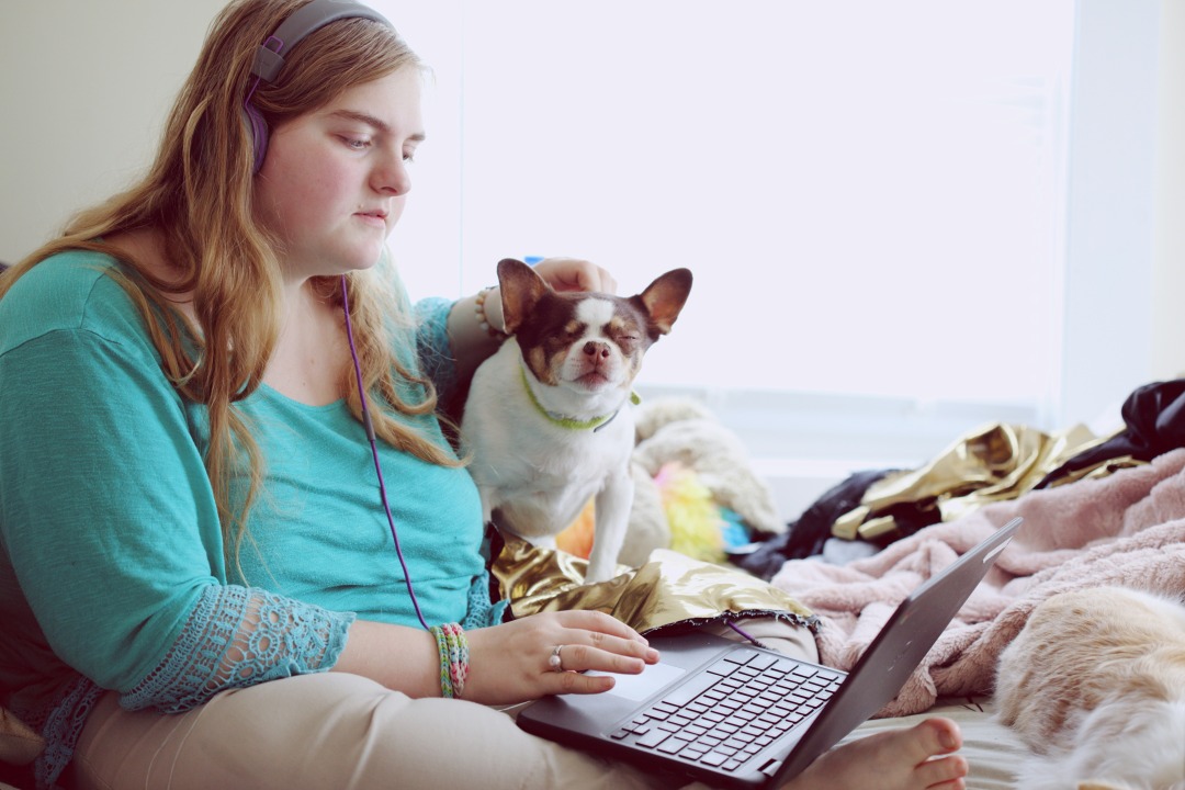 A girl sits on her bed with her dog and a laptop