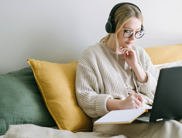 A woman sitting with headphones on writing in a notepad while watching a webinar